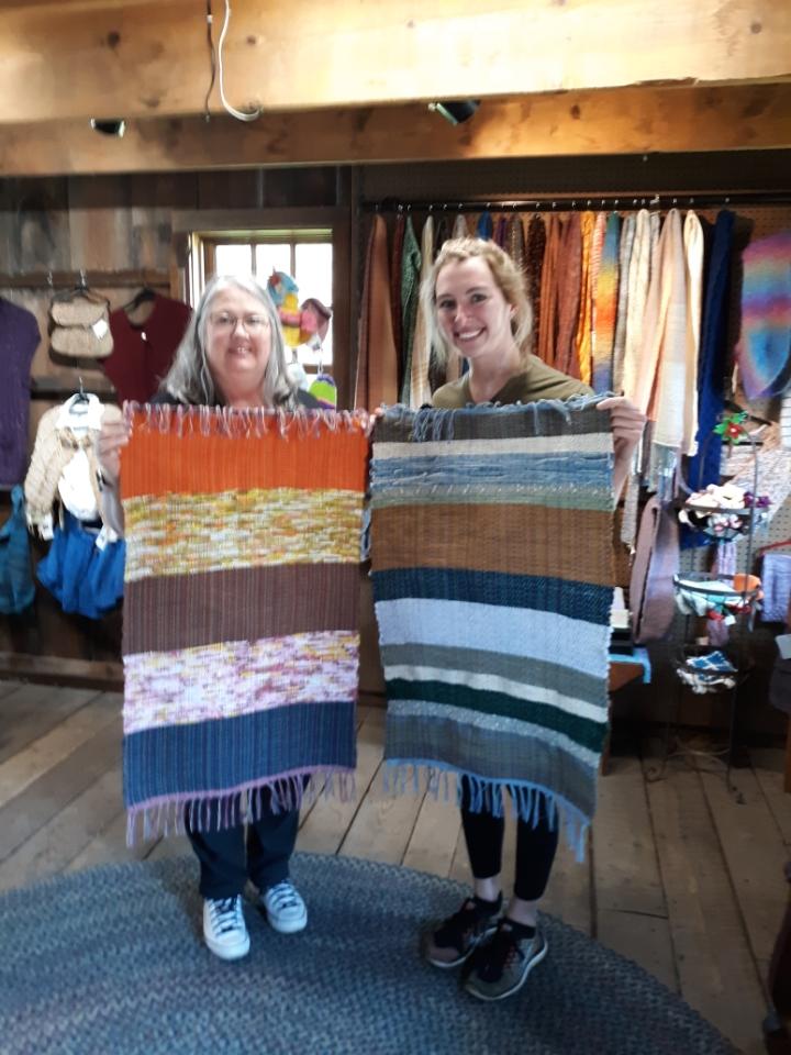 Two women holding up a woven rug in front of some clothes.